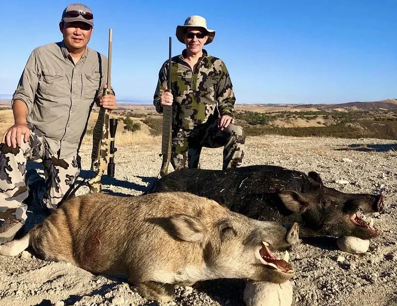 Two hunters kneeling with wild pigs on a Camp Five Outfitters ranch under a blue sky